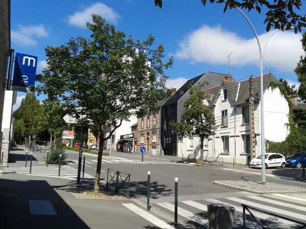 Vista de una calle con un paso de peatones, un letrero azul con una 'm' blanca, y edificios con una mezcla de arquitectura moderna y tradicional.