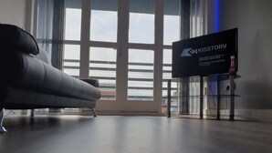 Ein modernes Zimmer mit einem großen Fenster, einem grauen Sofa und einem Bildschirm, auf dem 'KISSSTORY' angezeigt wird.