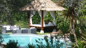 Un área junto a la piscina con un pabellón de techo de paja, asientos blancos acolchados y una piscina azul cristalina.