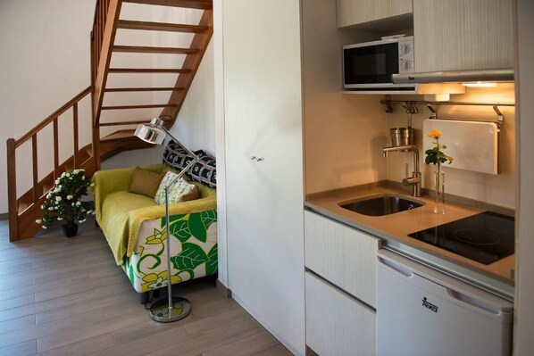 A compact kitchenette with a microwave, sink, and a small refrigerator. A wooden staircase leads to a bed with a green and white floral bedspread.