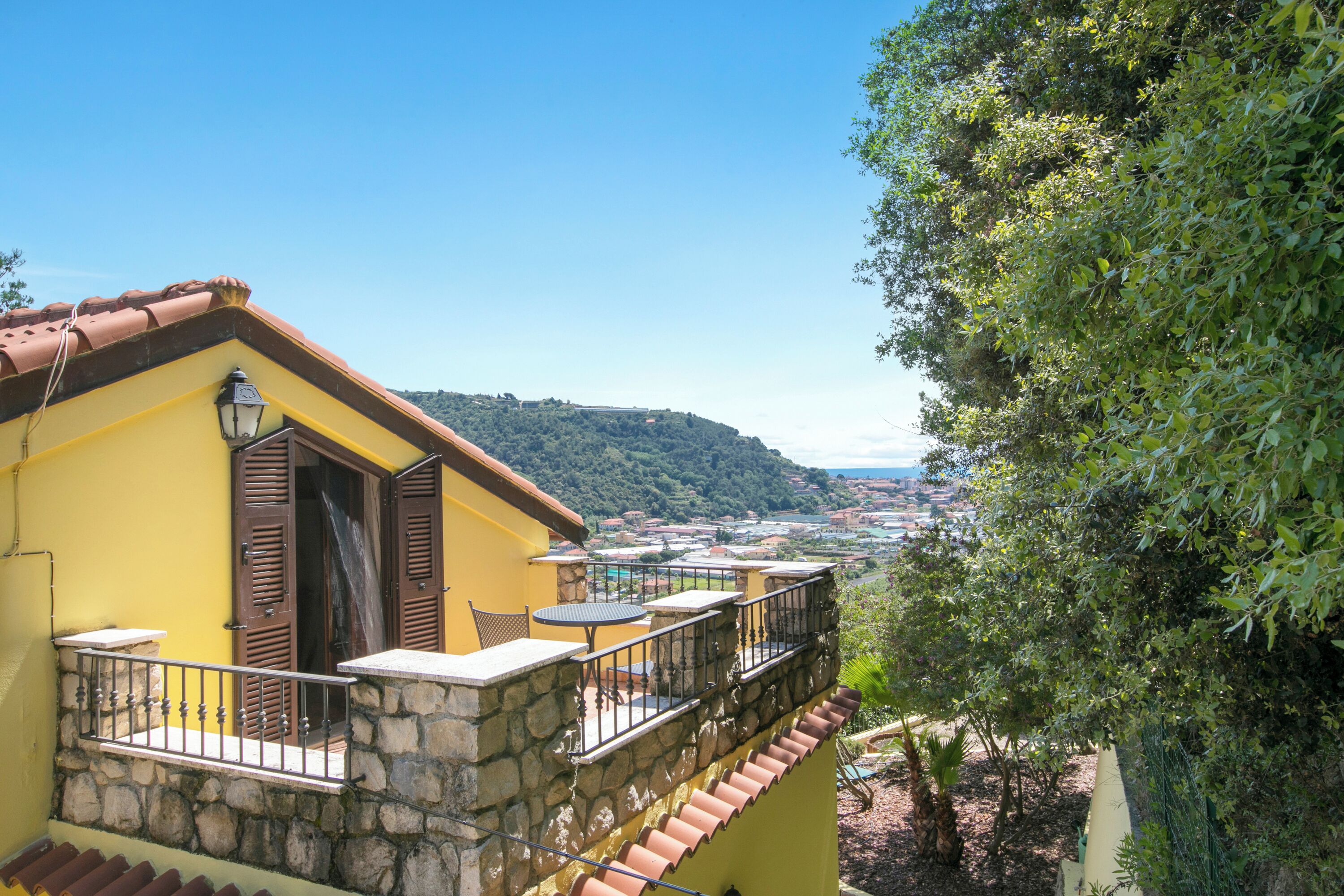 Un bâtiment jaune avec un balcon en pierre, une vue sur une ville au loin et un arbre sur la droite.