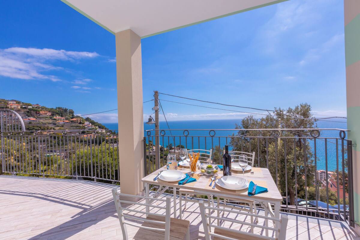 Un balcón con mesa lista para comer, con vistas a un pueblo costero y al mar.