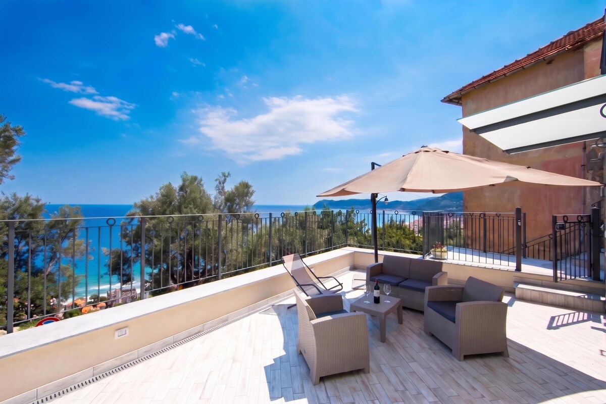 A terrace with outdoor furniture, a large umbrella, and a view of the sea.