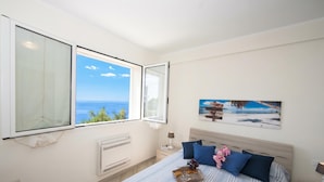 A bedroom with a large window, a bed with blue and white striped bedding, a nightstand with a lamp, and a framed picture on the wall. - Serena 2 (Alassio)
