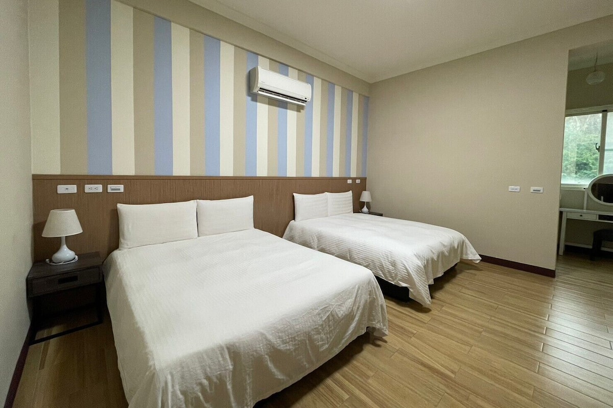 A hotel room with two beds, a wooden headboard, a striped wall, an air conditioning unit, a desk, and a window.