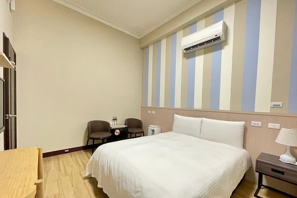 A hotel room with a large bed, a desk, two chairs, an air conditioning unit, and a striped wall.