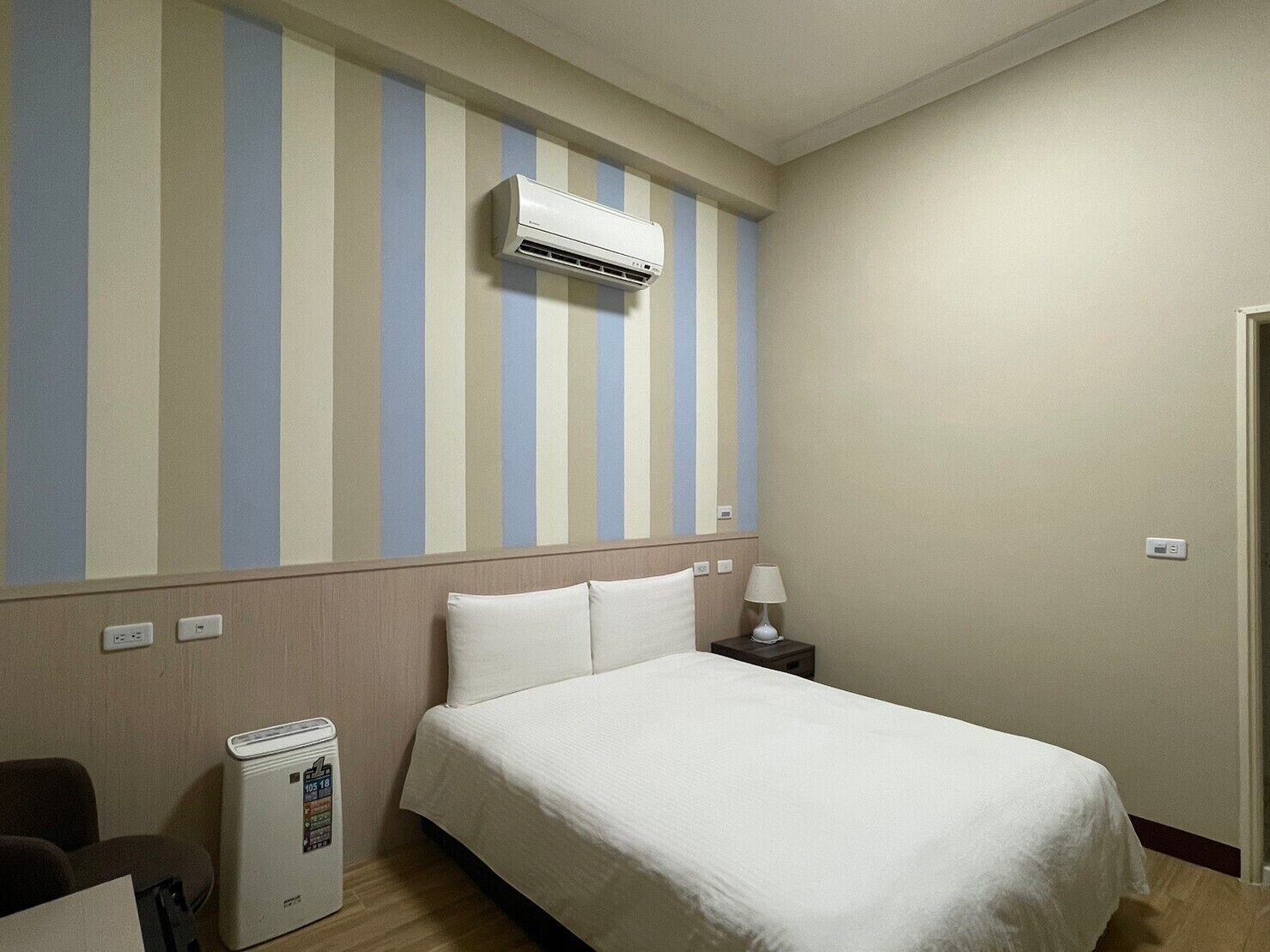 A hotel room with a bed, a nightstand, a lamp, an air conditioner, and a heater.