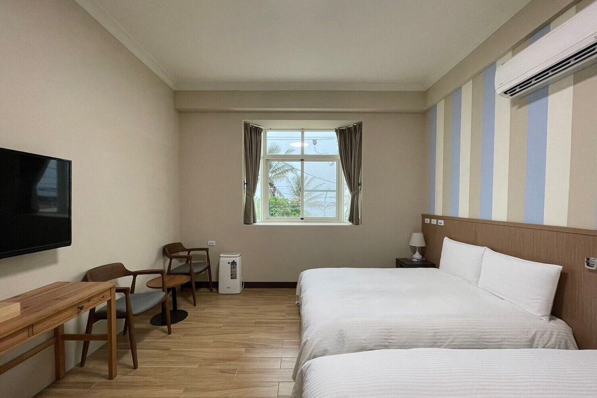 A hotel room with two beds, a desk with a chair, a television, a window with curtains, and a striped wall.