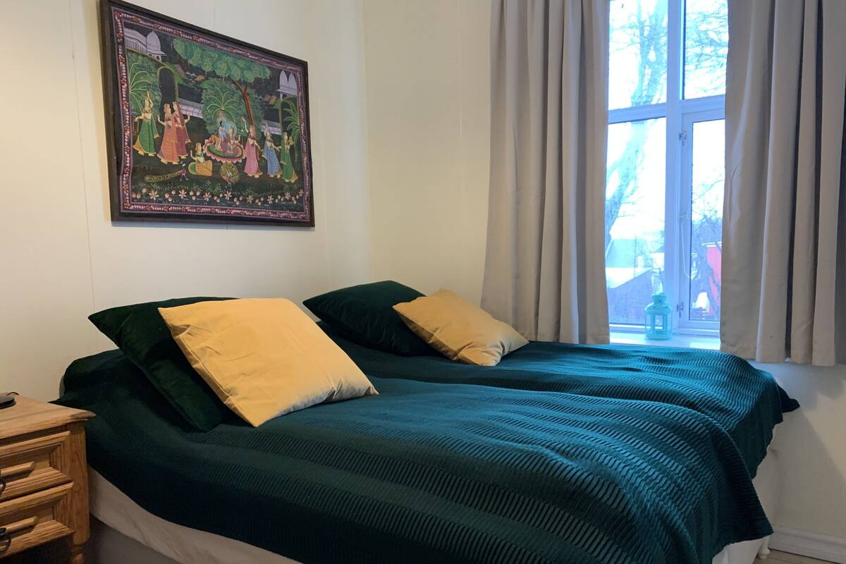 A room with two beds, a wooden nightstand, a framed painting, and a window with curtains.