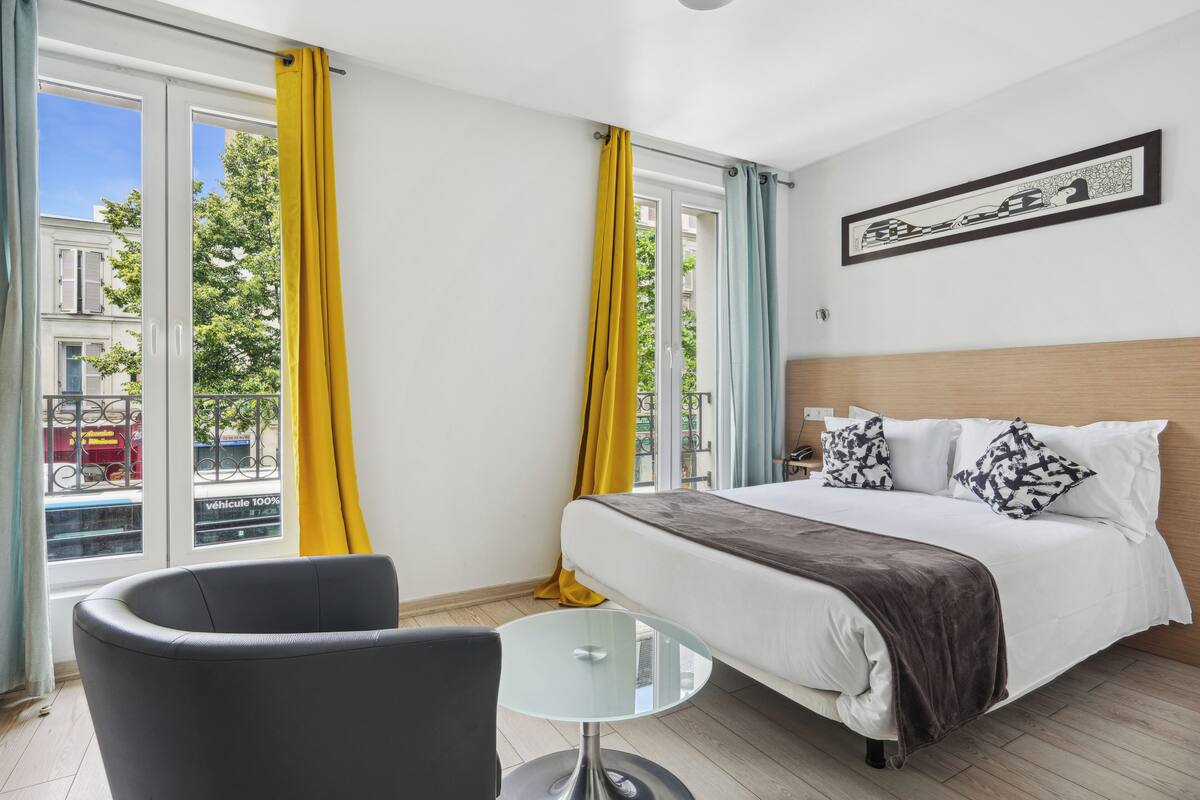 A hotel room with a bed, a glass table, a chair, and a view of the street outside.