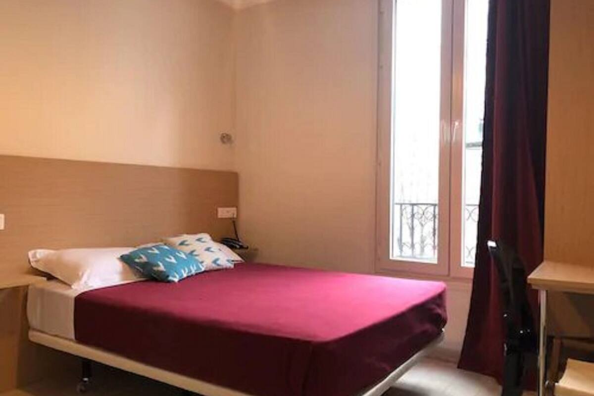 A single bed with a red bedspread, a white pillow, and a blue pillow with a leaf pattern. There is a small table with a chair next to the bed, and a window with a curtain.