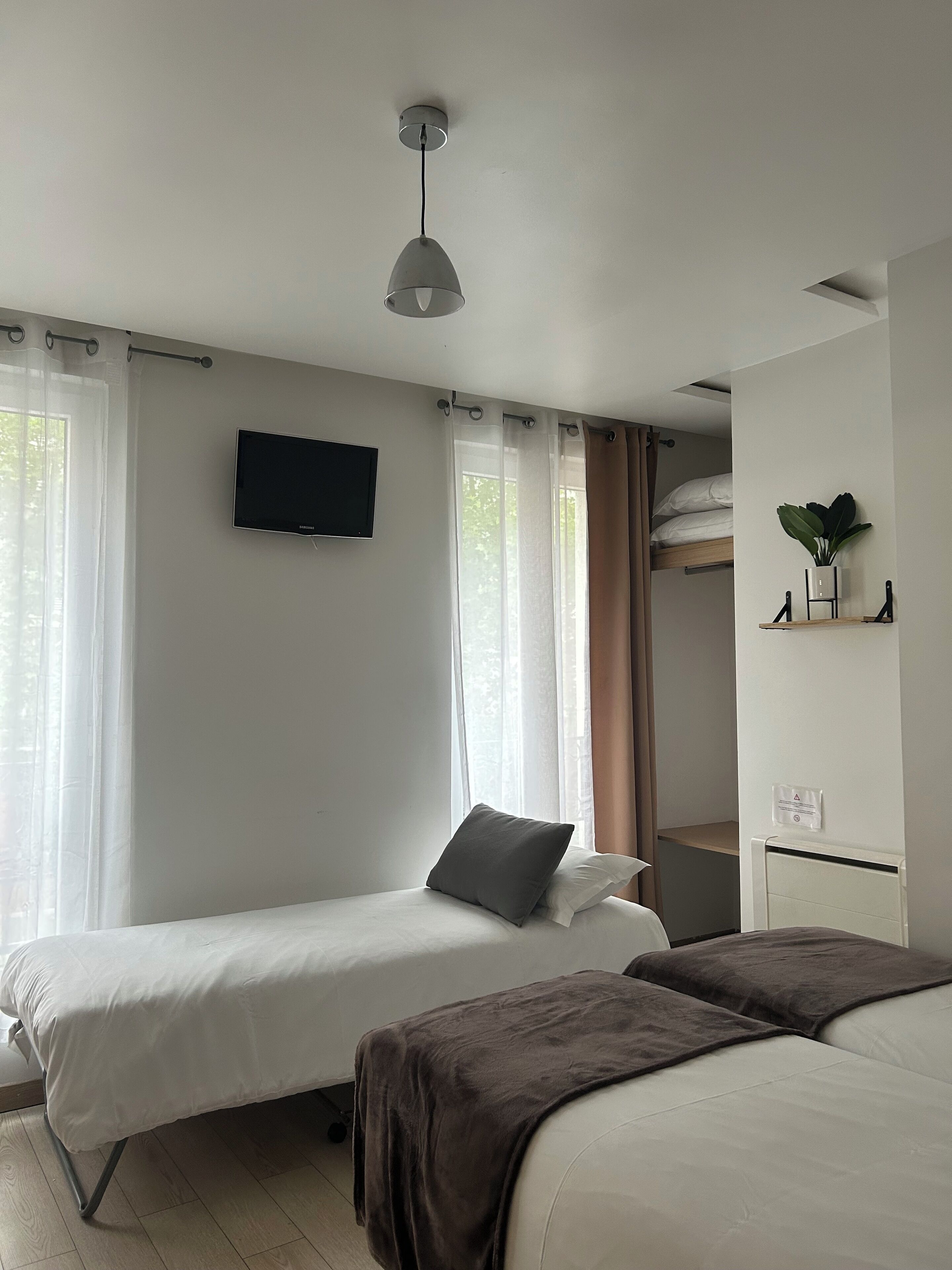 Een slaapkamer met twee bedden, een aan de muur bevestigde televisie, een kamerplantje en een raam met gordijnen.