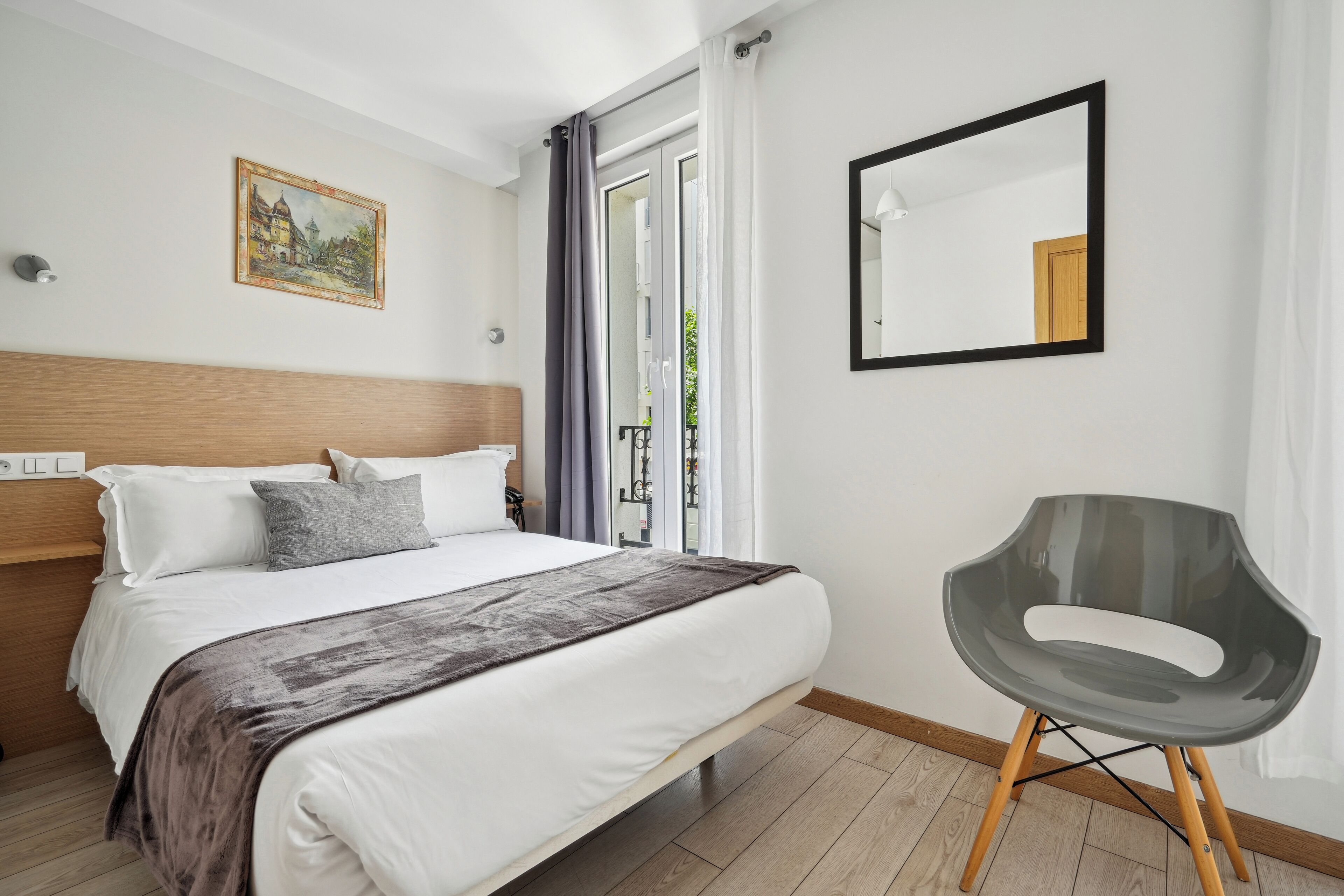 A modern hotel room with a large bed, a grey chair, a painting on the wall, and a window with curtains.