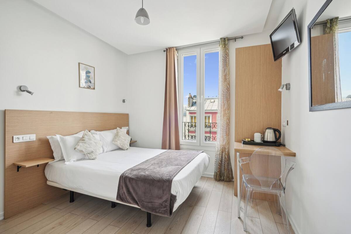 A modern bedroom with a wooden bed, a bedside table, a TV mounted on the wall, and a view of buildings through the window.