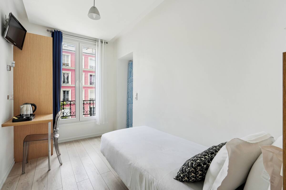 A bedroom with a bed, a desk with a kettle, a chair, a TV mounted on the wall, and a window with curtains.