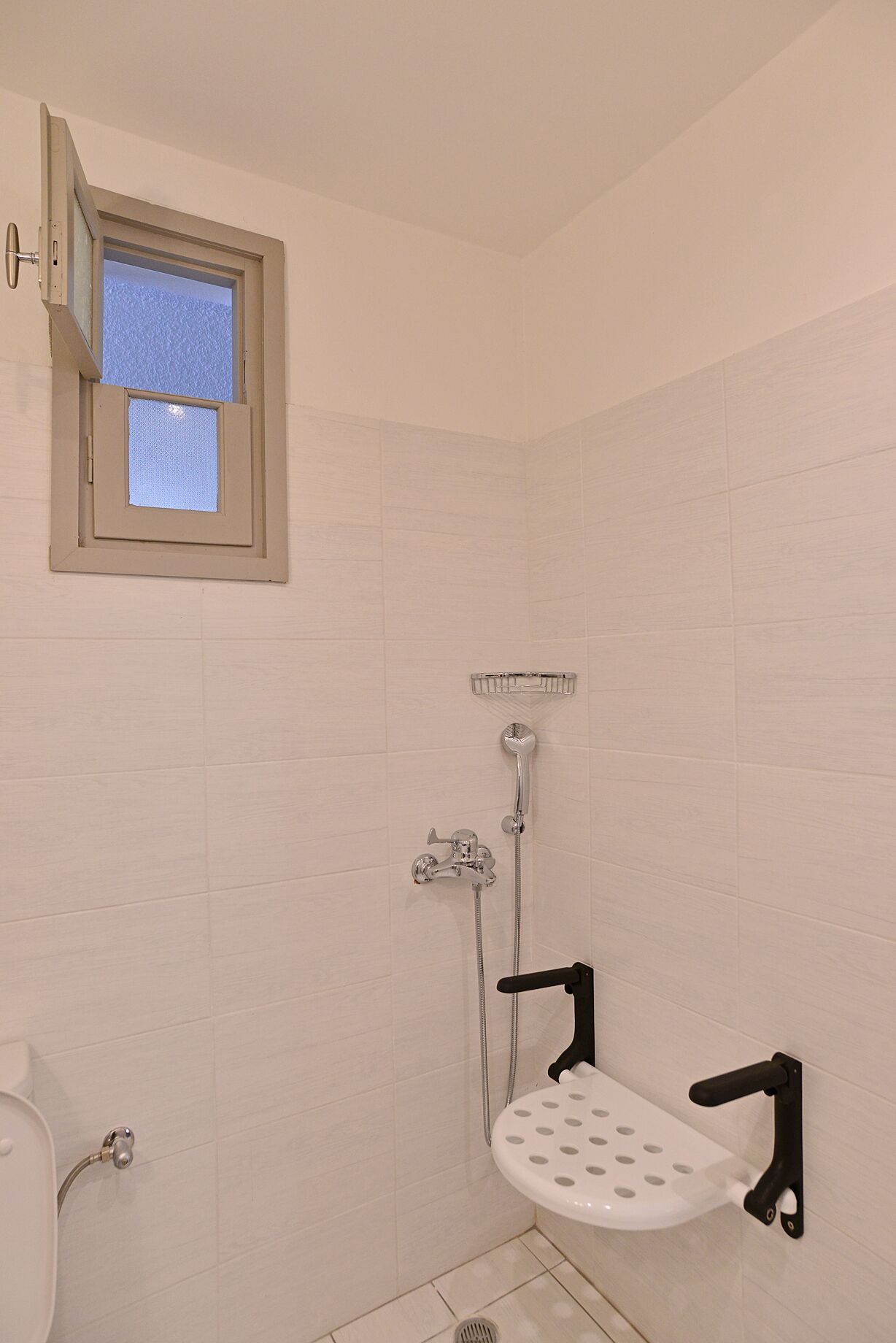 A small, enclosed shower area with a window, a handheld showerhead, a handheld shower attachment, and a white toilet.