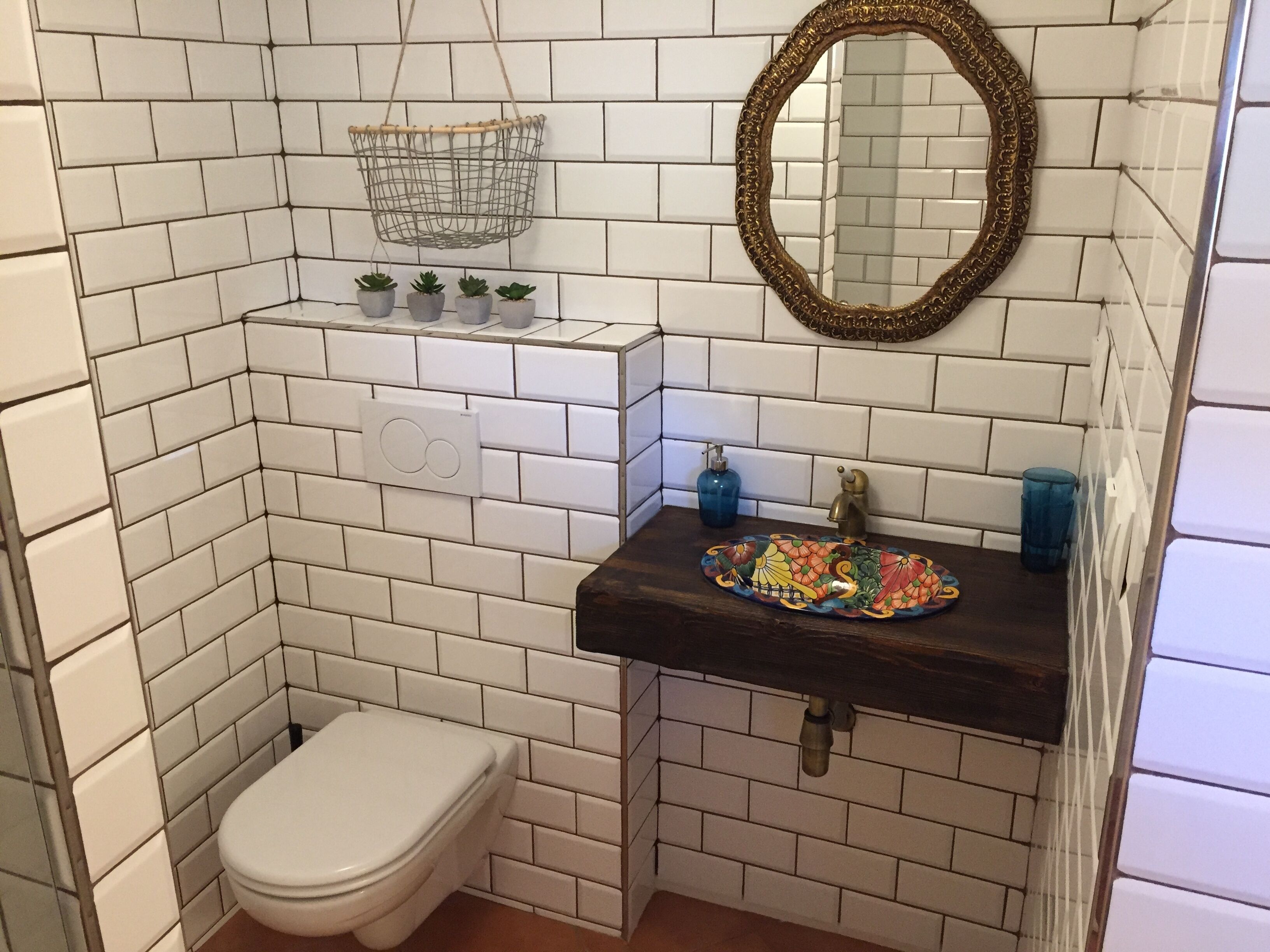 A bathroom with white tiled walls, a white toilet, a dark sink with a decorative tile backsplash, a hanging metal basket, and a mirror with an ornate frame.