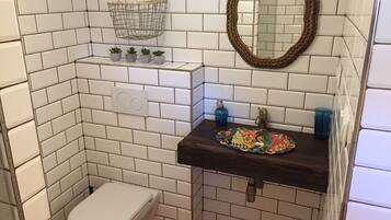 A bathroom with white tiled walls, a white toilet, a dark sink with a decorative tile backsplash, a hanging metal basket, and a mirror with an ornate frame.