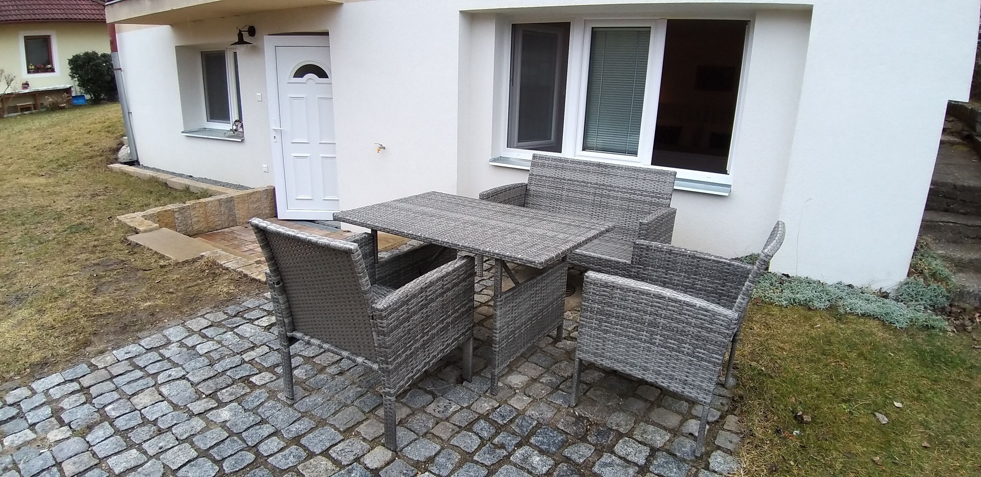 Een terras buiten met een tafel en vier rieten stoelen, een witte deur en een raam met zonwering.