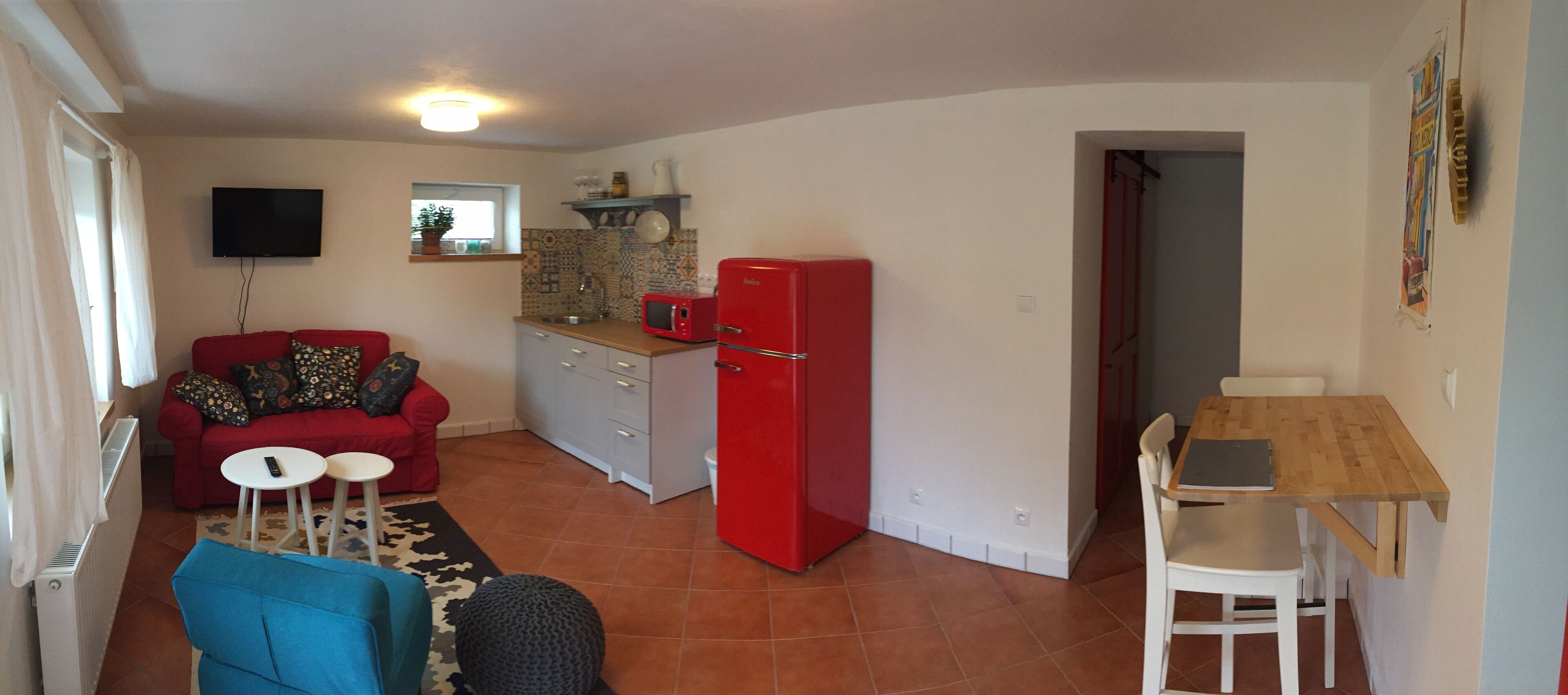 A compact living space with a red refrigerator, a small kitchen area, a dining table with chairs, a red sofa with patterned cushions, and a television mounted on the wall.