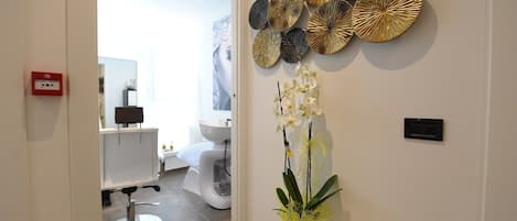 Un baño moderno con un lavamanos, inodoro y espejo blancos. Hay una obra de arte decorativa sobre el lavamanos y una planta de orquídea en maceta en el suelo.