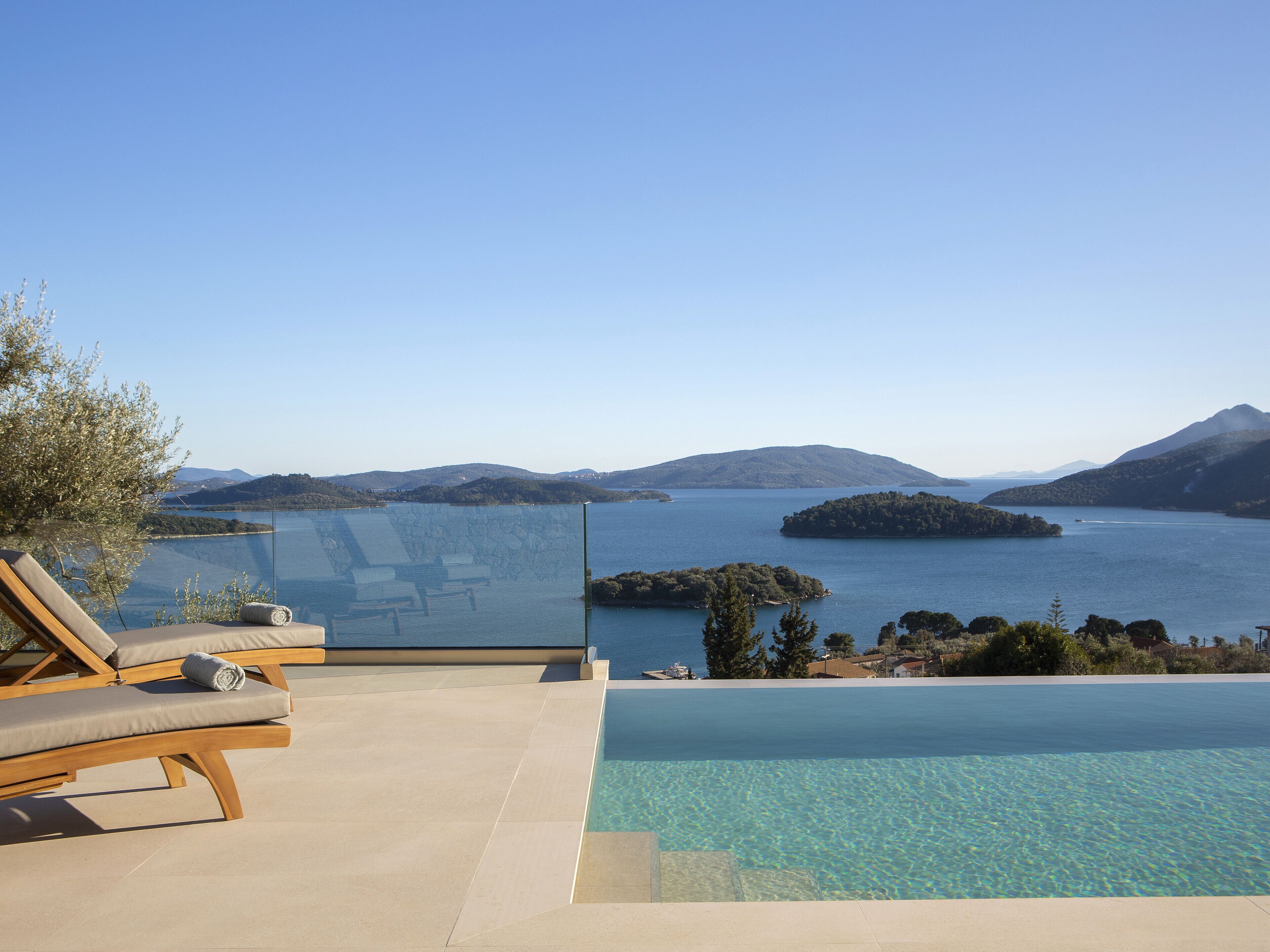 An infinity pool with clear water, wooden lounge chairs, and a view of a large body of water with islands and mountains in the distance.