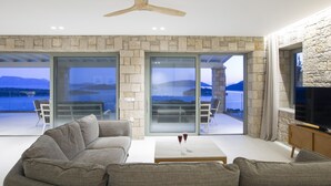 A modern living room with a stone wall, a ceiling fan, a sofa, a coffee table, and a TV. - Elegant stone-built Villa, with heated pool and amazing view (Lefkada)