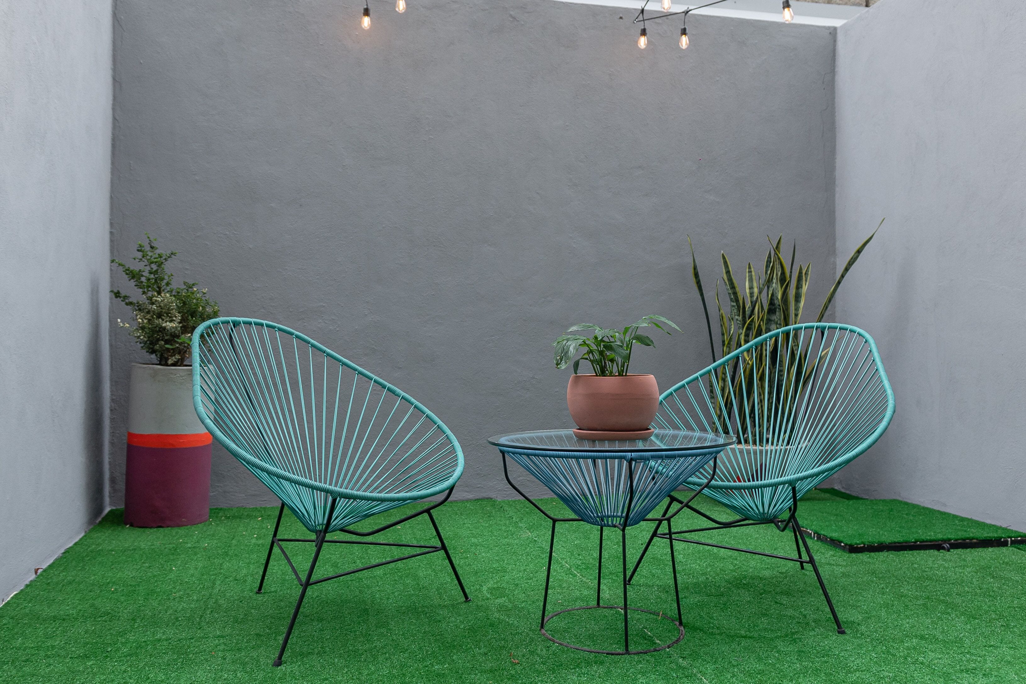 Dos sillas de alambre turquesa, una pequeña mesa redonda y una planta en maceta en una esquina con césped artificial verde y una pared gris.