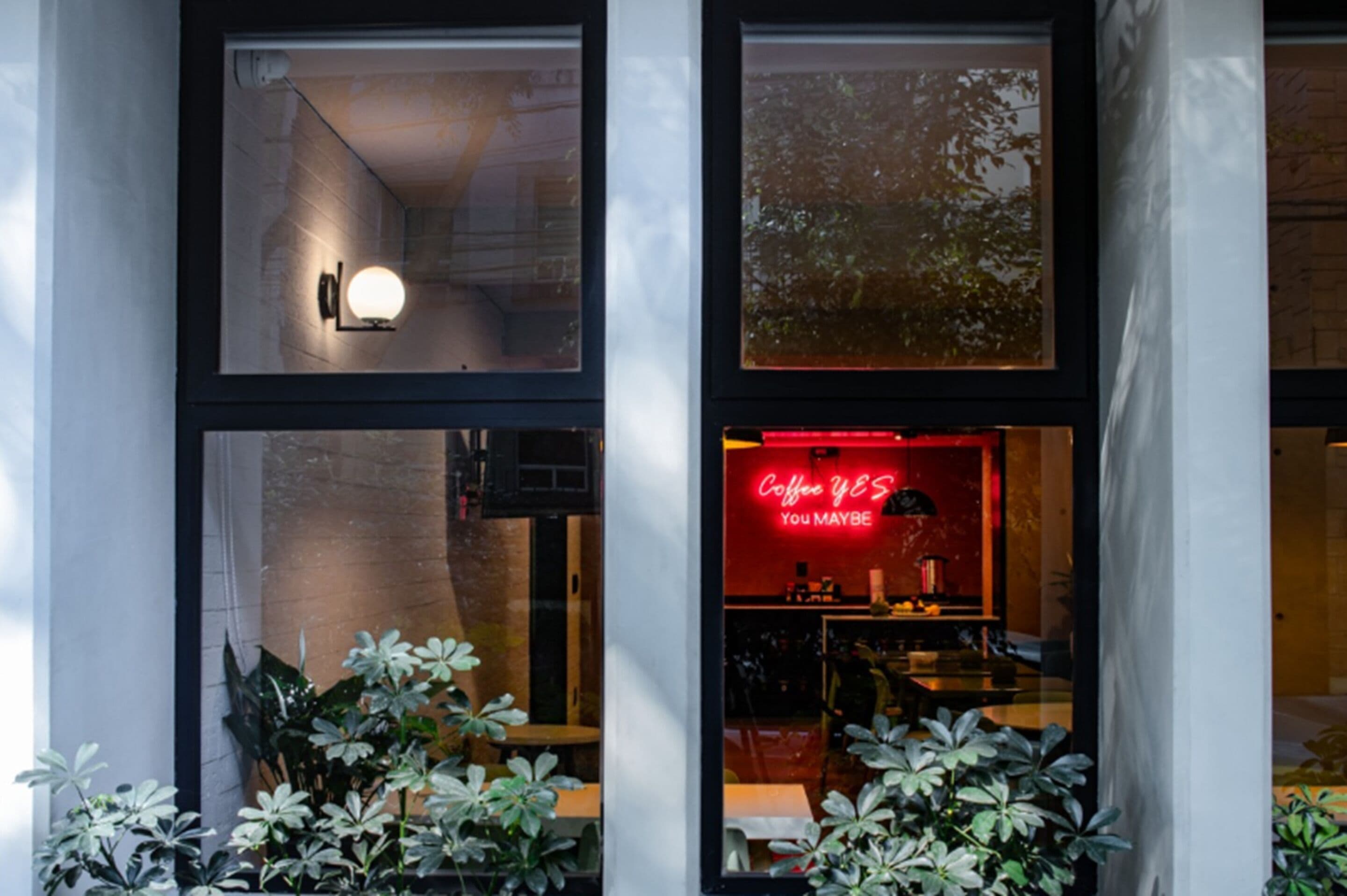 Een café met een neonbord, grote ramen en binnenshuis planten.