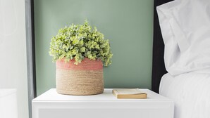 A white bedside table with a woven basket containing green foliage, a book, and a white pillow on a bed with wooden headboard. - NIU Modern & Centric Lofts (Mexico City)