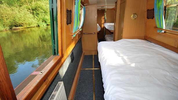 A narrow, enclosed cabin with a bed, a small window, and a view of a river. - Floating Holidays (Stockport)