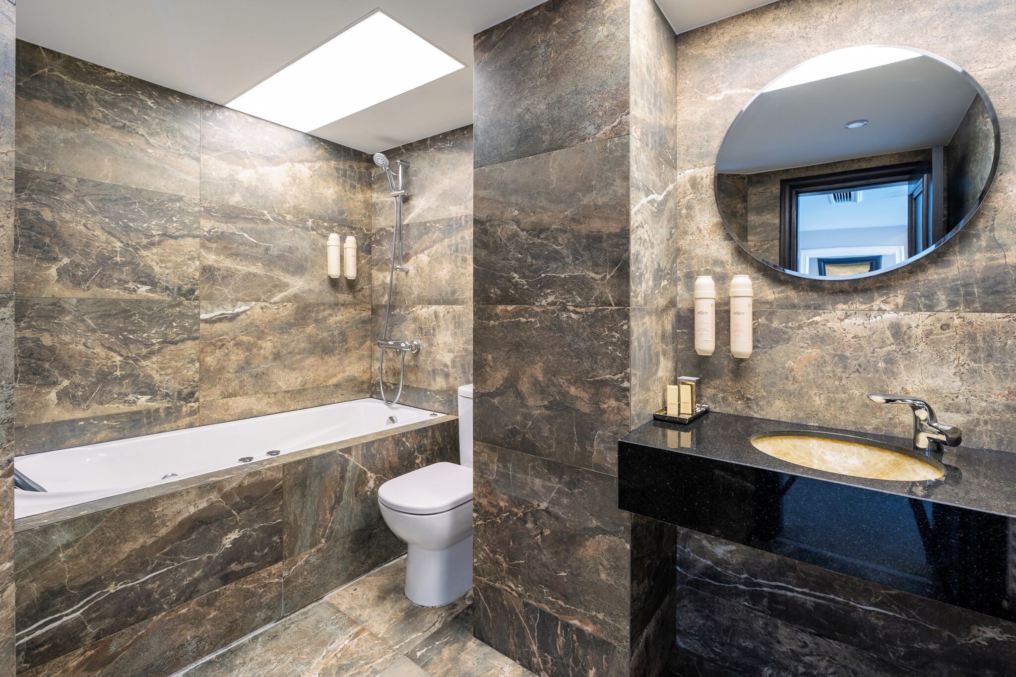 Un baño moderno con bañera, inodoro y lavamanos, que cuenta con paredes de mármol y un tragaluz.