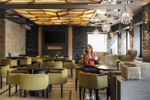 A modern interior with a woman seated at a table, surrounded by yellow chairs, a large screen, and a patterned ceiling. - Hotel Tsarevets (Veliko Tarnovo)
