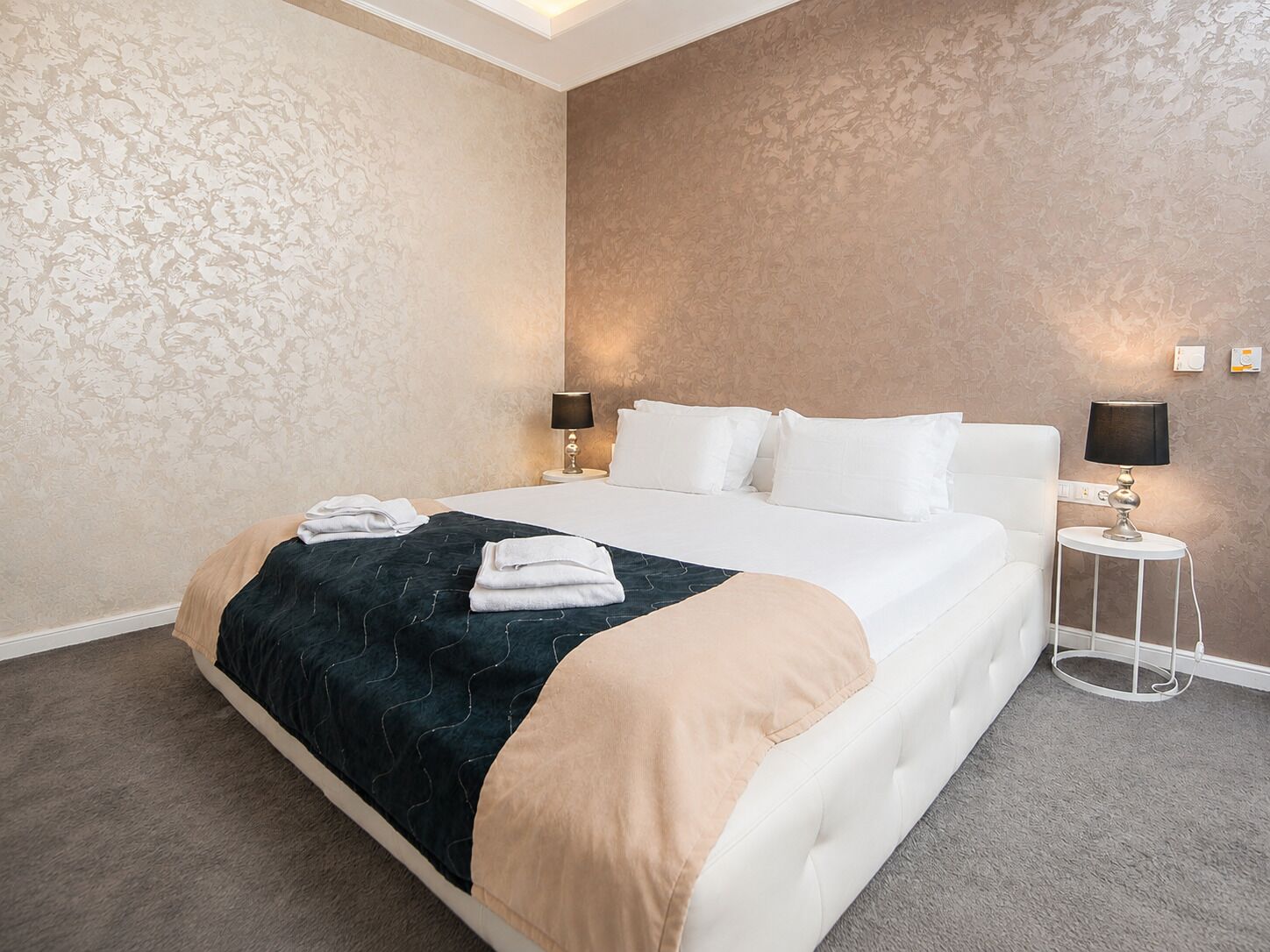 A hotel room with a large bed, two bedside tables with lamps, a carpeted floor, and patterned wallpaper.