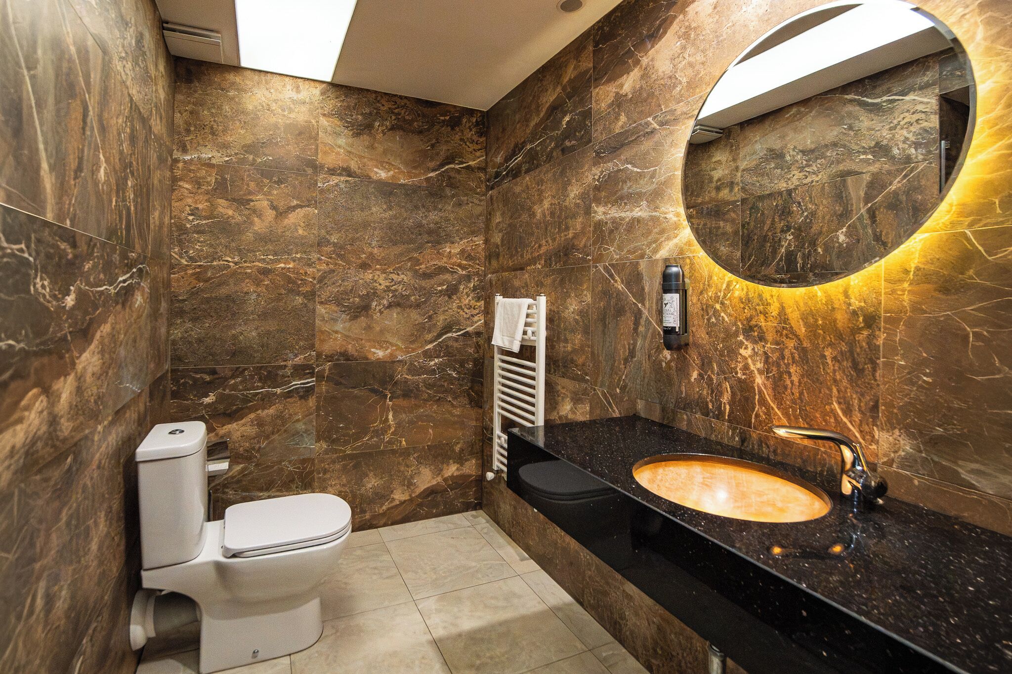 Et moderne badeværelse med en marmorvæg, en sort bordplade, et rundt spejl og et toilet.