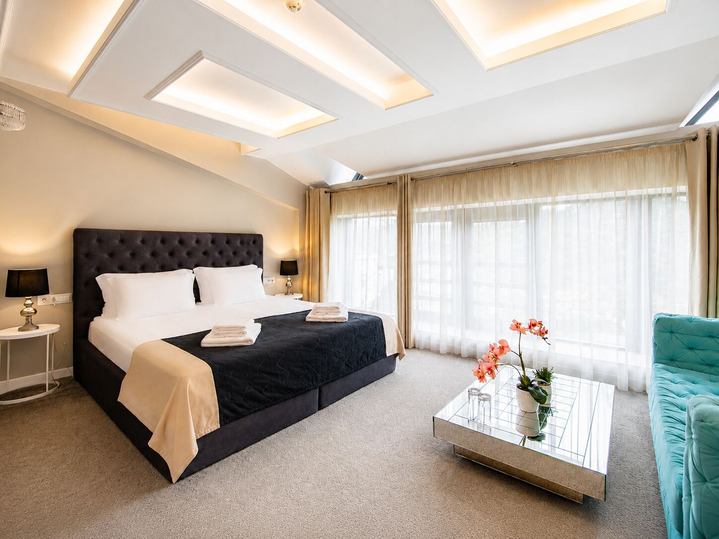 A hotel room with a large bed, bedside lamps, a coffee table with flowers, and a teal armchair.