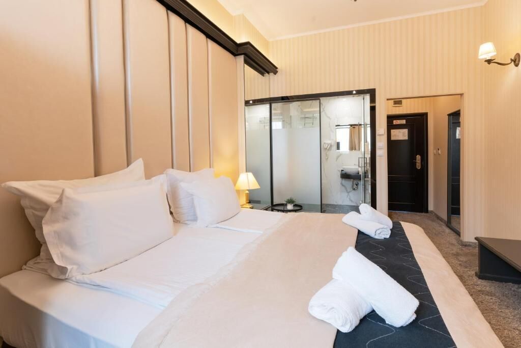 Une chambre d’hôtel avec un grand lit, une table de chevet avec une lampe, une salle de bain visible à travers une porte en verre et un banc de couleur sombre.