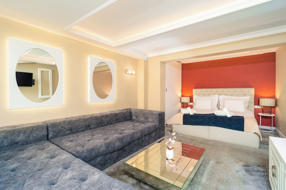 A modern hotel room with a sectional sofa, a glass coffee table, a bed with a red accent wall, and two bedside lamps.