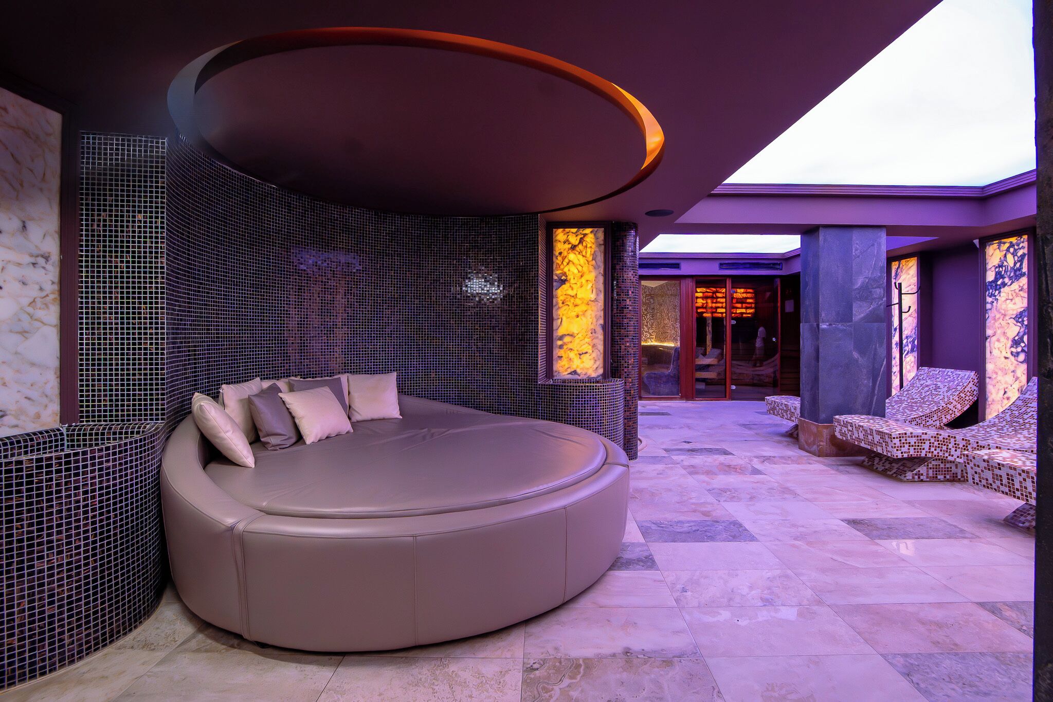 A modern spa room with a circular seating area, a mosaic wall, and a view of the spa entrance.