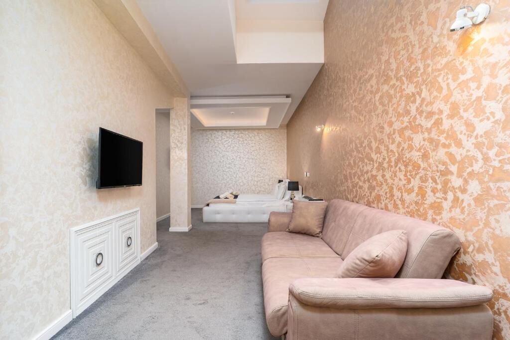 A room with a flat-screen TV mounted on the wall, a beige sofa, a white bed with a headboard, and a patterned wall.