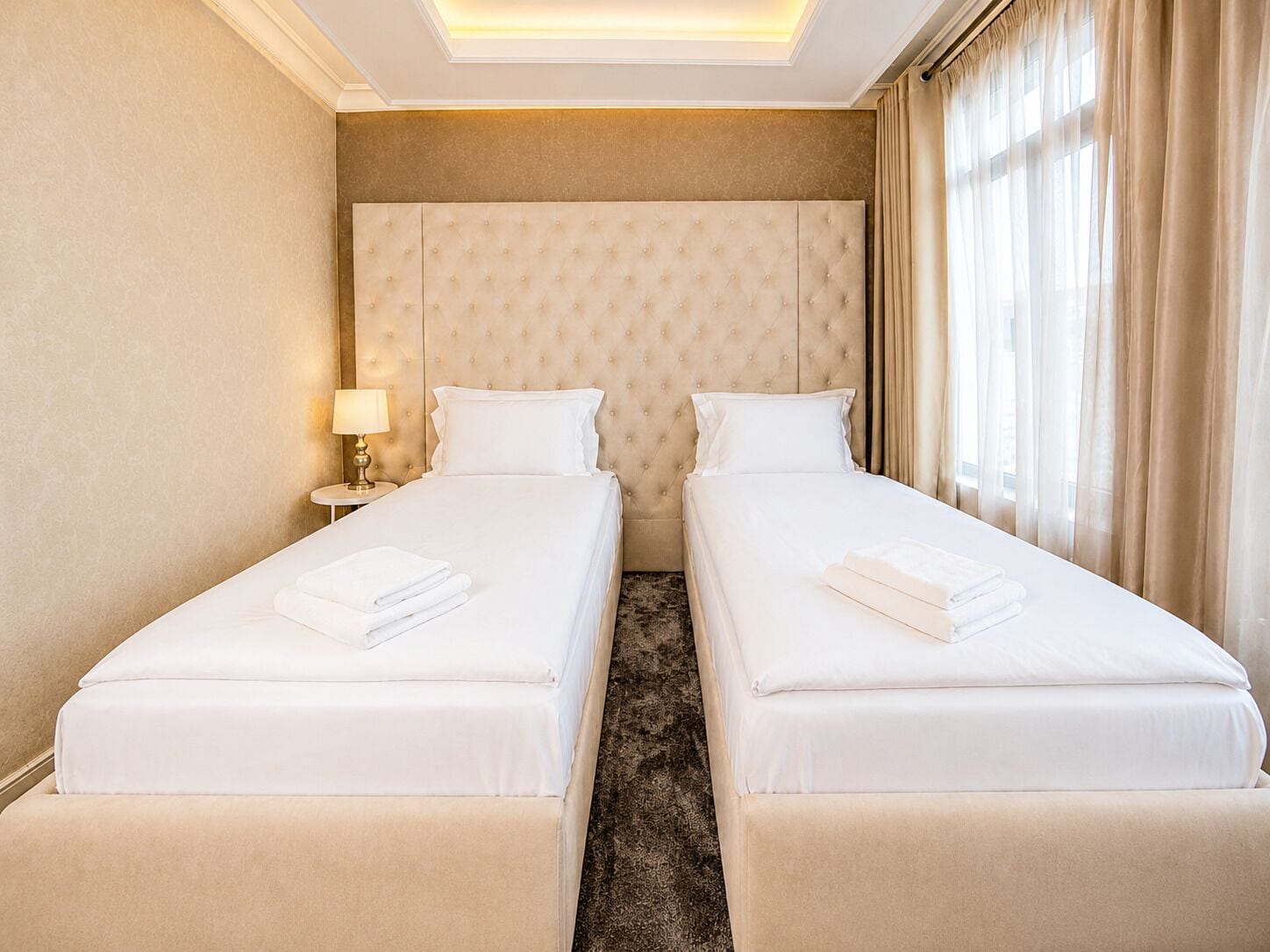 A hotel room with two beds, a headboard, a nightstand with a lamp, and a window with curtains.