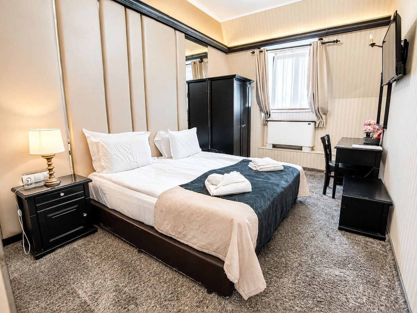 Habitación de hotel con cama, mesita de noche, lámpara, armario, escritorio, silla y ventana con cortinas.