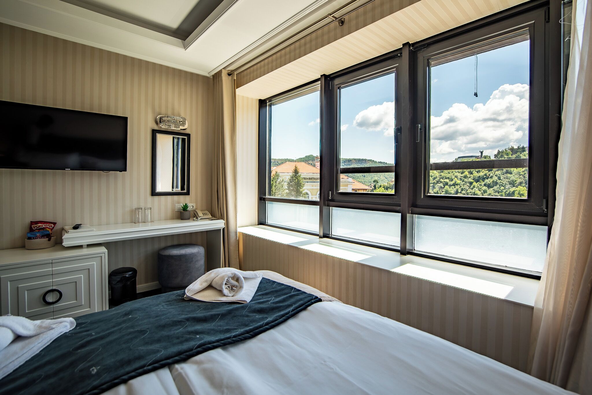 Habitación de hotel con cama, televisor montado en la pared, ventana con vistas a un edificio y árboles, y mesita de noche con lámpara y teléfono.