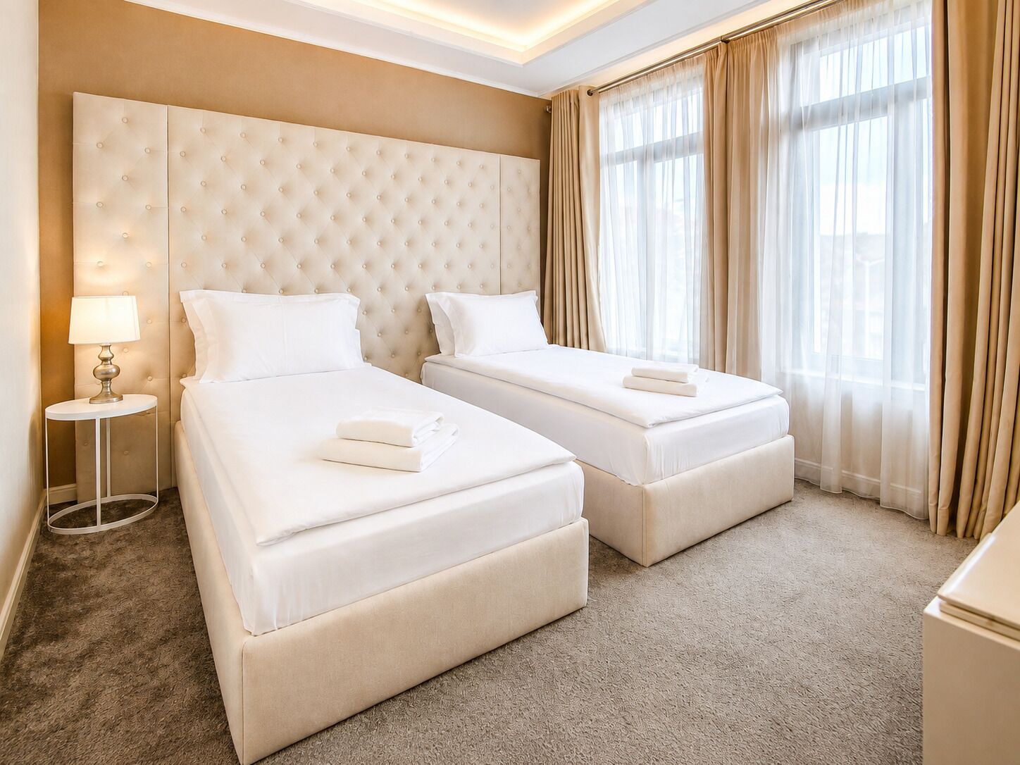 A hotel room with two beds, a round side table, a lamp, and a large window with curtains.