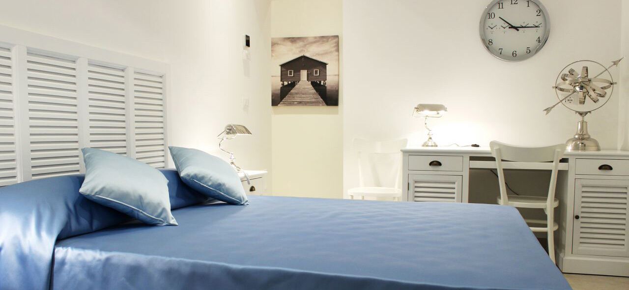 Een slaapkamer met een bed, kussens, een bureau, een stoel, een klok en een aan de muur hangend schilderij.