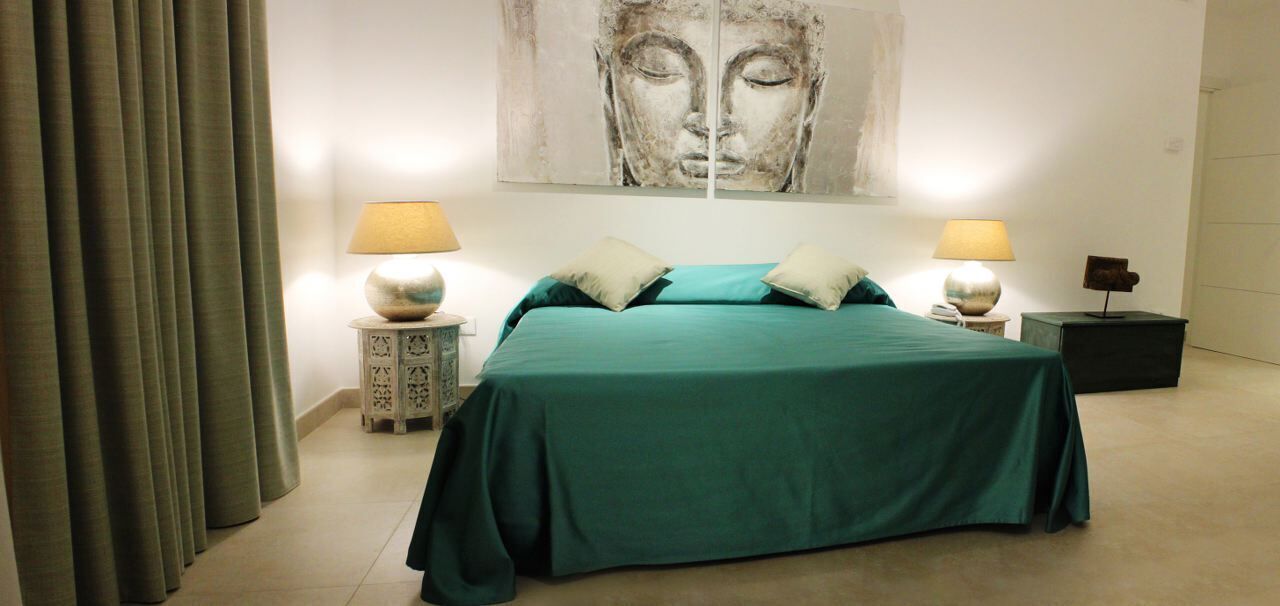 Ein Schlafzimmer mit einem Bett, zwei Nachttischen mit Lampen, einem großen abstrakten Gemälde an der Wand und einem Vorhang auf der linken Seite.