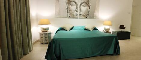 A bedroom with a bed, two bedside tables with lamps, a large abstract painting on the wall, and a curtain on the left.