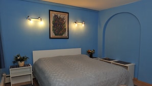 Ein Schlafzimmer mit einem Bett, einem Nachttisch mit Lampe, einem gerahmten Bild an der Wand und einem kleinen Tisch mit Blumenvase.