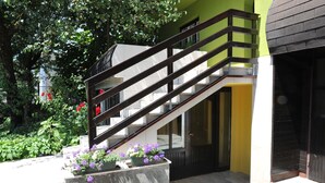 Eine Treppe mit dunkler Geländer führt zu einer gelben Tür, umgeben von Grünpflanzen und Blumen.