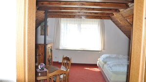 Una habitación con techo inclinado, una cama, un ventanal con cortinas translúcidas, una mesa de madera con sillas y un radiador.